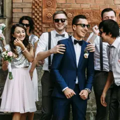 Uśmiechnięci druhny i drużbowie pozują do zdjęcia. Dziewczyny w błyszczących topach i różowych spódniczkach trzymają bukieciki. Panowie w garniturach i z szelkami wyglądają elegancko.