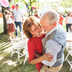 Uśmiechnięta para seniorów tańczy na przyjęciu w ogrodzie, w tle ludzie i dekoracje.