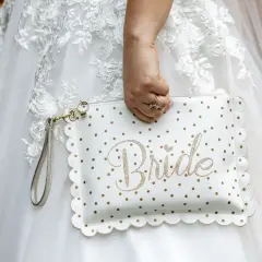 Biała kopertówka w złote kropki z napisem "Bride" to niezbędnik panny młodej. Dopełnia suknię ślubną.