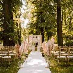 Piękny ślub w plenerze boho. Złote krzesła, biały dywan i trawy pampasowe tworzą magiczną atmosferę wśród drzew.