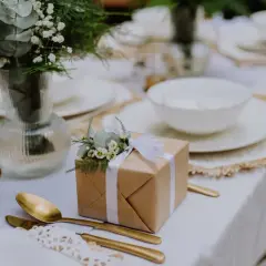 Eleganckie upominki na wesele, ozdobione zielenią i białymi kwiatami, czekają na gości przy nakrytym stole.