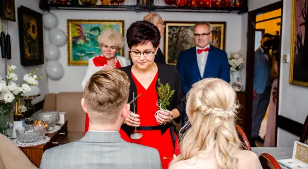 Kobieta udziela katolickie błogosławieństwo młodej parze, trzymając gałązkę i krzyż.