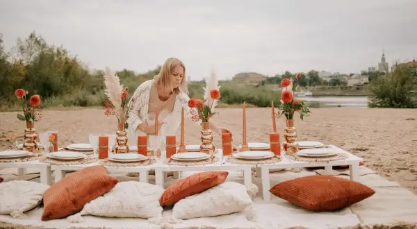 Kobieta przygotowuje romantyczny piknik nad rzeką, idealny na organizację wieczoru panieńskiego. Pomarańczowe kwiaty i świece tworzą ciepłą atmosferę.