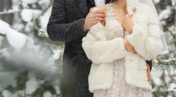 Para młoda w zimowej scenerii. Ona w białym futerku i koronkowej sukni, on w eleganckim płaszczu. Ich ślub w zimie wygląda bajkowo.