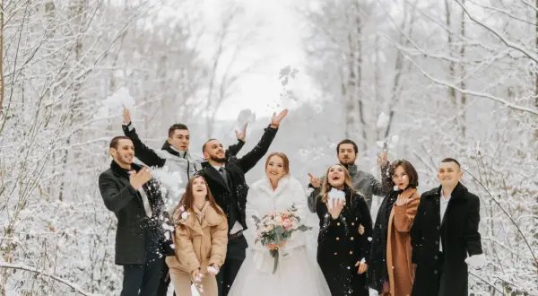 Radosna gromadka rzuca śnieżkami w zimowym lesie. Panna młoda w białym futerku i pan młody w eleganckim płaszczu, otoczeni przez przyjaciół w stylowych okryciach, tworzą piękne stylizacje na wesele zimą.