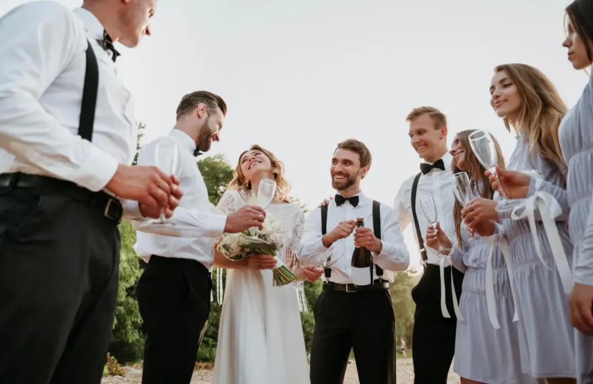 Świadek na weselu otwiera szampana, podczas gdy panna młoda i pan młody cieszą się chwilą z przyjaciółmi.
