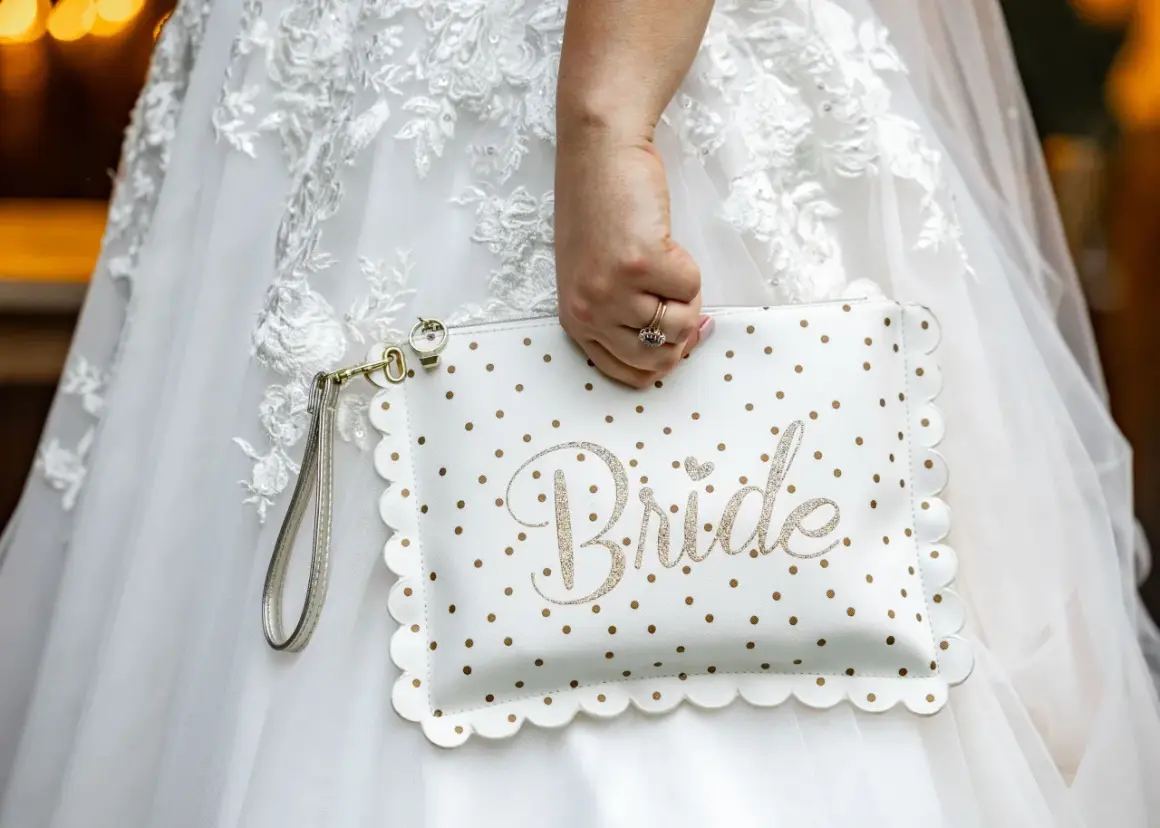 Biała kopertówka w złote kropki z napisem "Bride" to niezbędnik panny młodej. Dopełnia suknię ślubną.