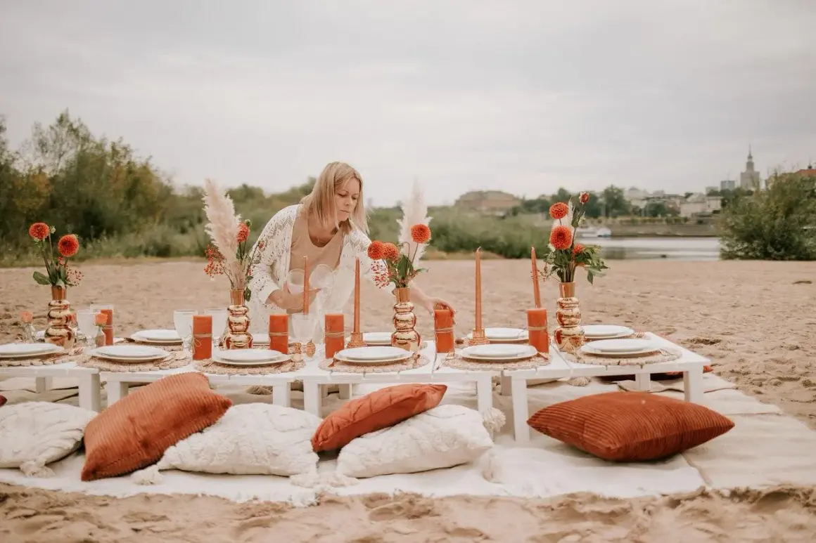 Kobieta przygotowuje romantyczny piknik nad rzeką, idealny na organizację wieczoru panieńskiego. Pomarańczowe kwiaty i świece tworzą ciepłą atmosferę.