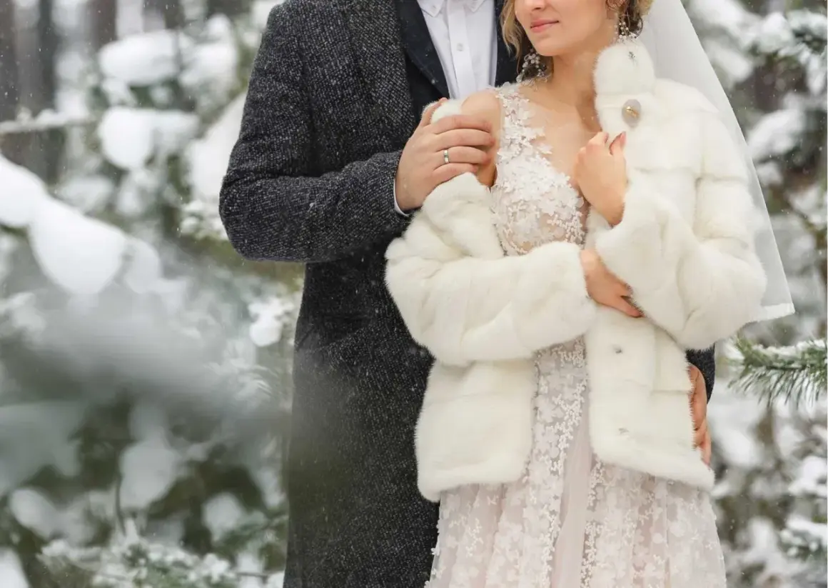 Para młoda w zimowej scenerii. Ona w białym futerku i koronkowej sukni, on w eleganckim płaszczu. Ich ślub w zimie wygląda bajkowo.
