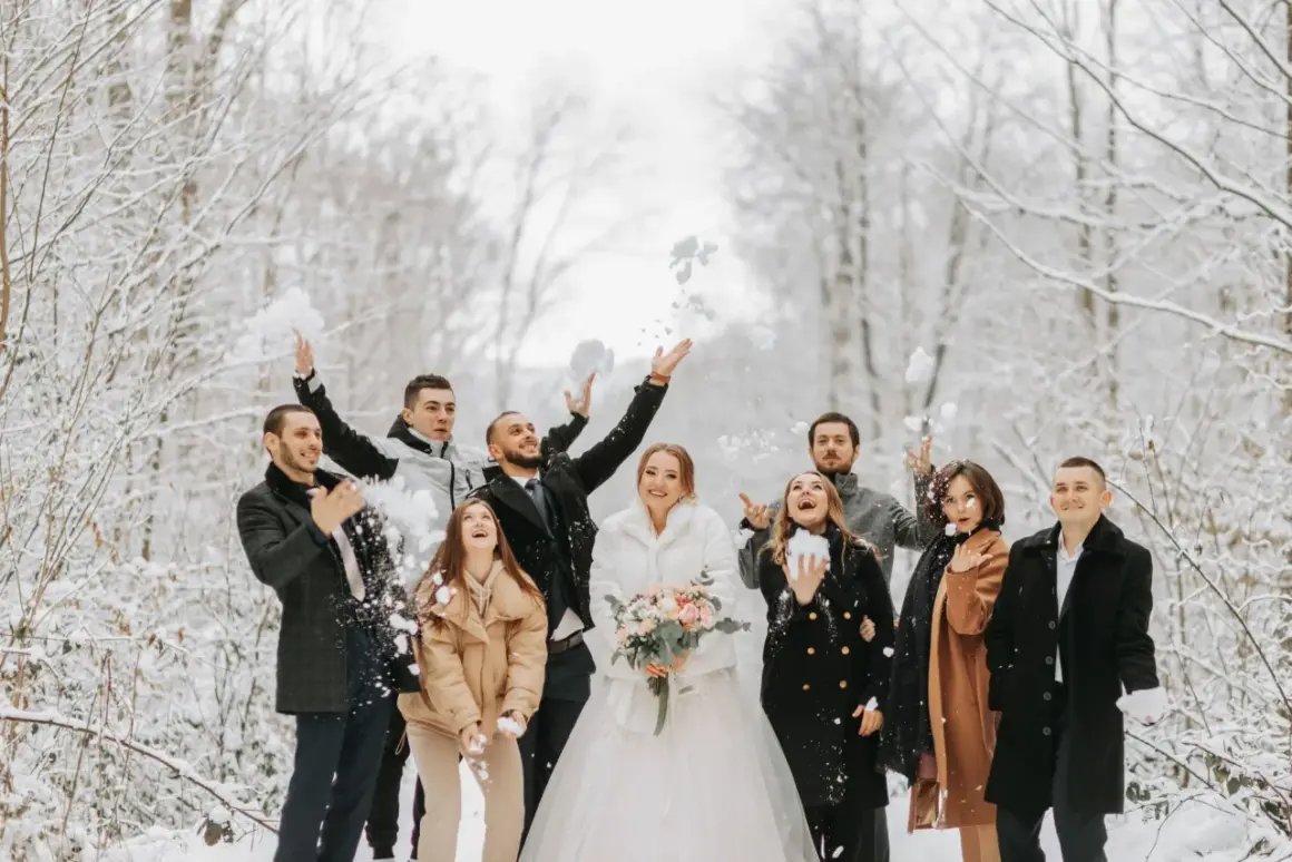 Radosna gromadka rzuca śnieżkami w zimowym lesie. Panna młoda w białym futerku i pan młody w eleganckim płaszczu, otoczeni przez przyjaciół w stylowych okryciach, tworzą piękne stylizacje na wesele zimą.