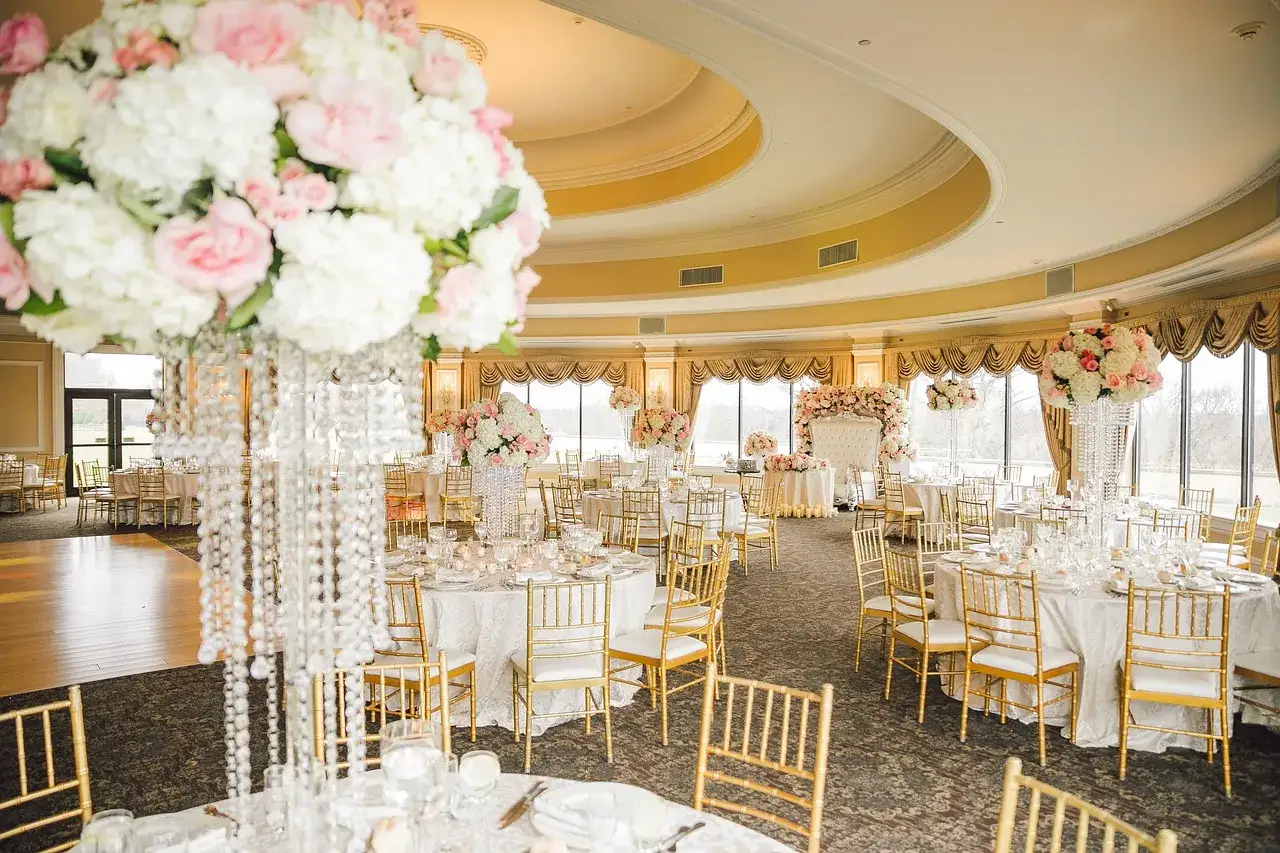 Elegancka sala weselna z białymi i różowymi kwiatami, kryształowymi żyrandolami i złotymi krzesłami. To idealne miejsce na samodzielny wystrój sali weselnej.