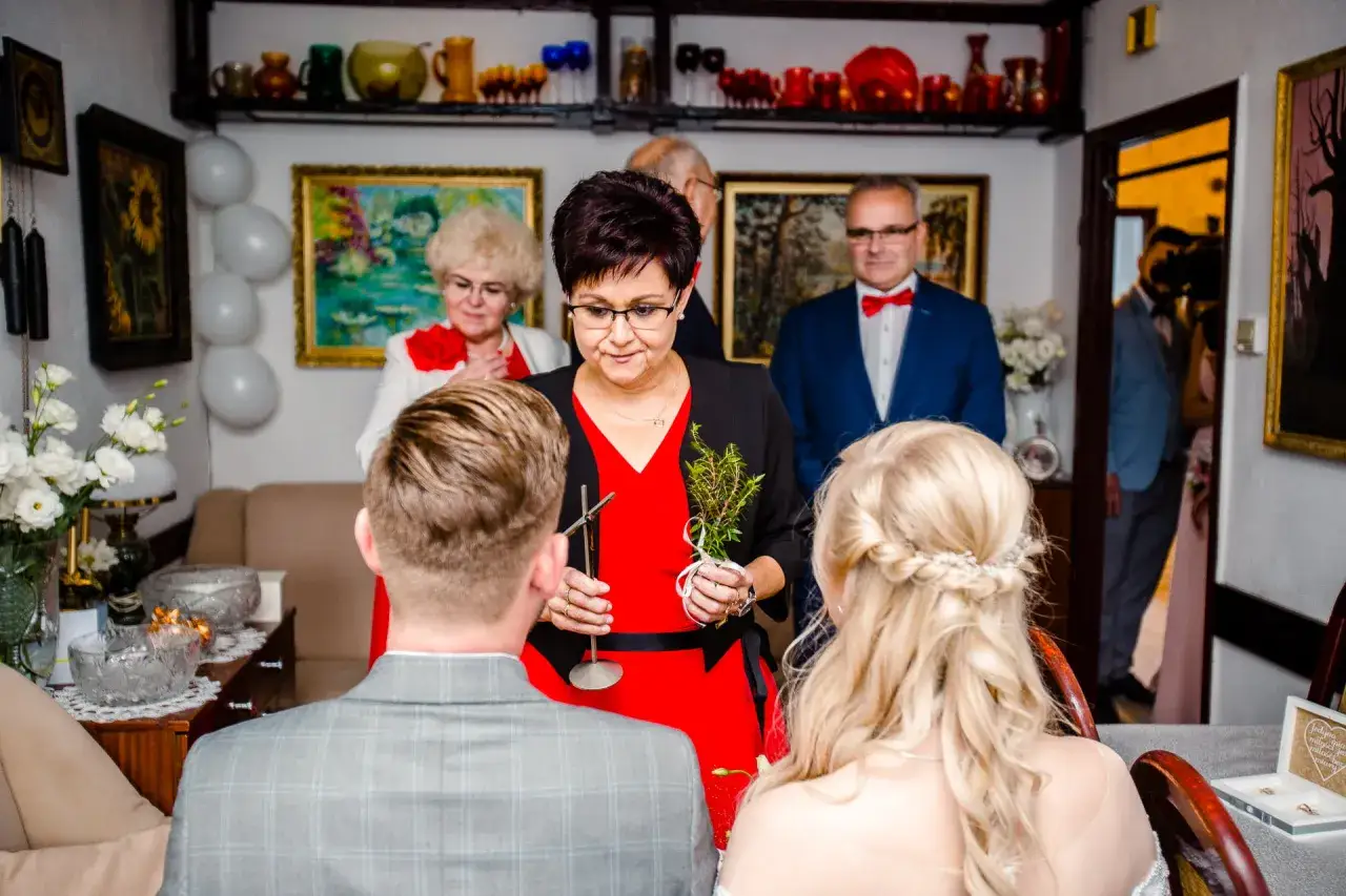 Kobieta z gałązką błogosławi parę przy stole.