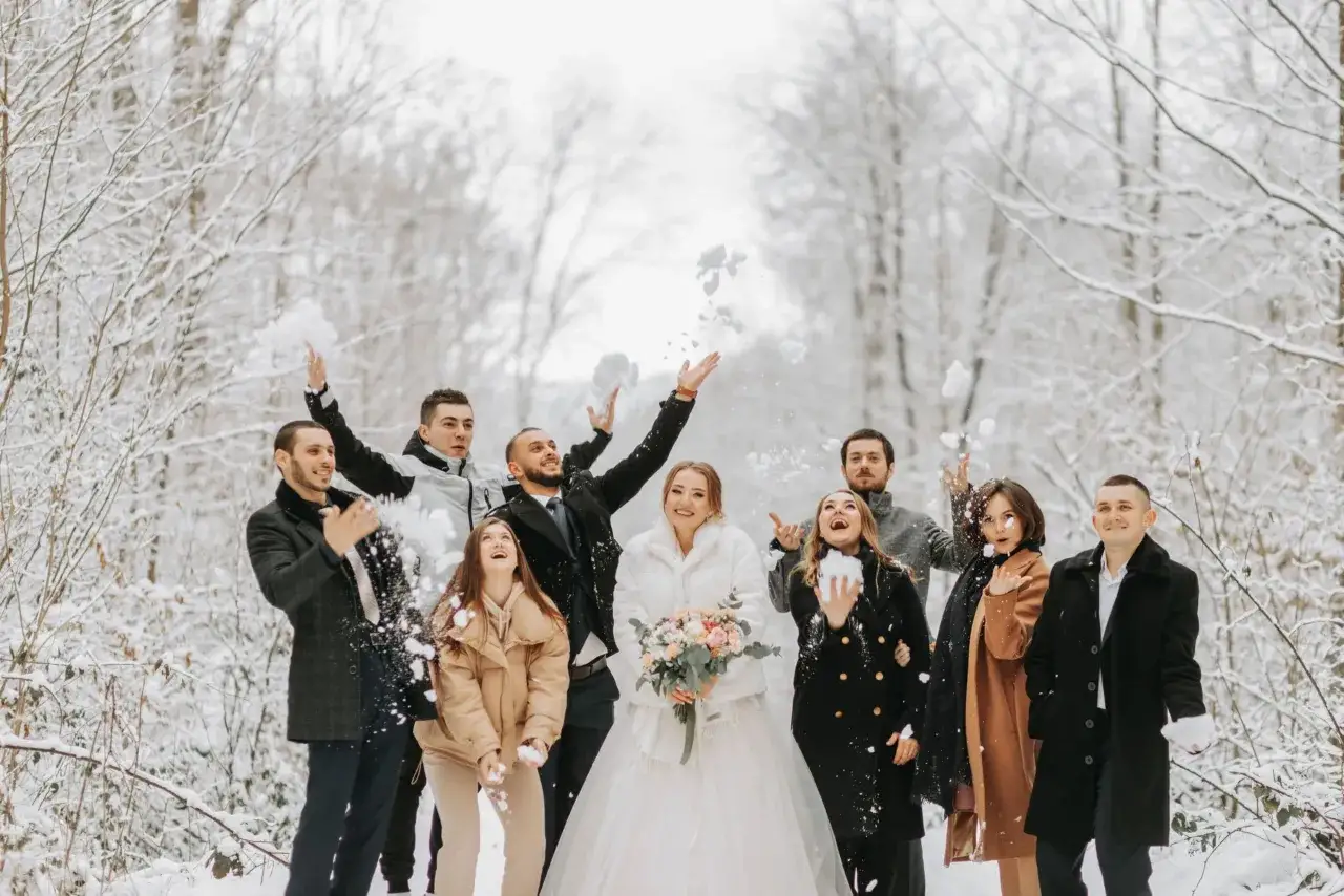 Radosna gromada rzuca śnieżkami w zimowym lesie. Panna młoda w białym futerku i pan młody w eleganckim płaszczu, otoczeni przez przyjaciół w stylowych okryciach, tworzą idealne stylizacje na wesele zimą.