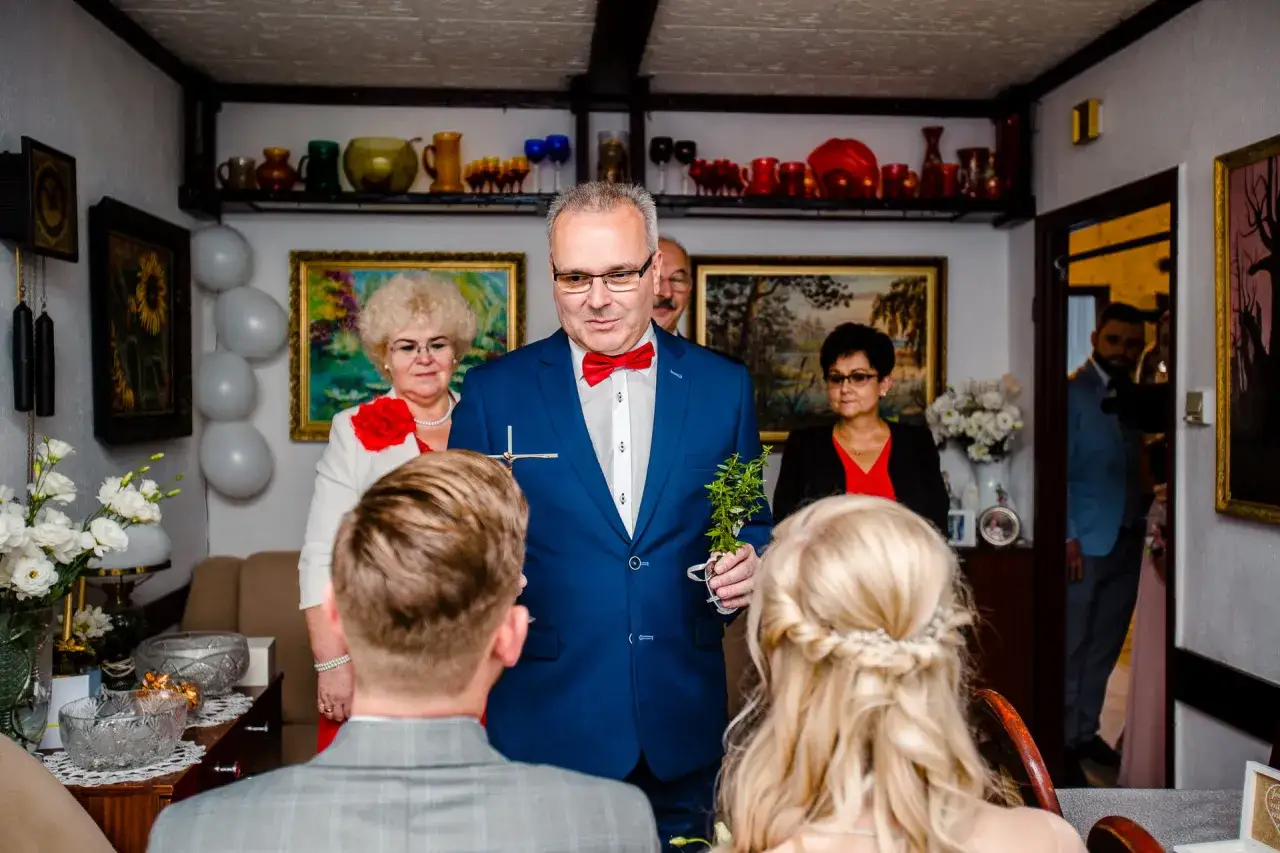 Uroczysty moment: katolickie błogosławieństwo młodej pary. Mężczyzna w garniturze błogosławi parę, trzymając krzyż i gałązkę.