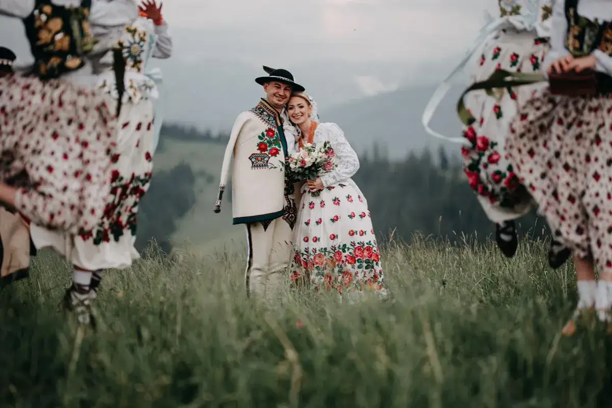 Młoda para w strojach ludowych, otoczona tańczącymi kobietami. Starosta na weselu wygląda radośnie, trzymając bukiet.