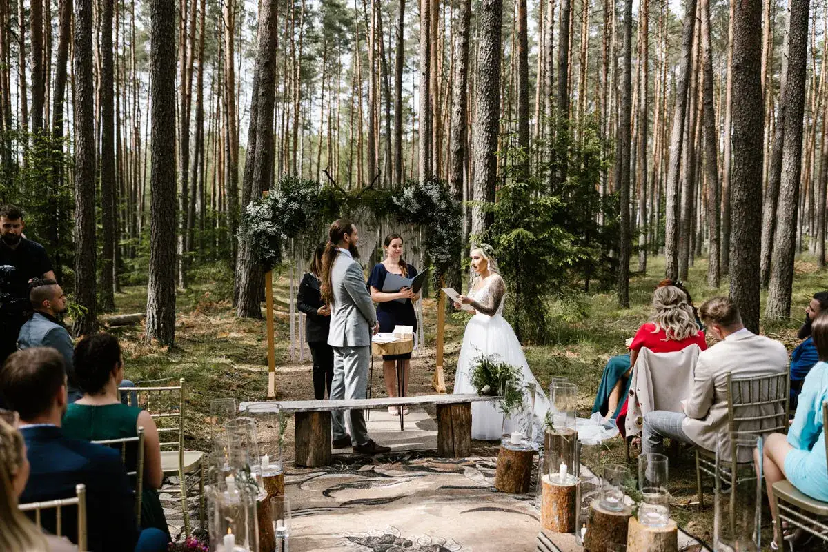 Uroczyste ślub w lesie. Para młoda stoi przed ołtarzem z zieleni, goście siedzą na krzesłach.