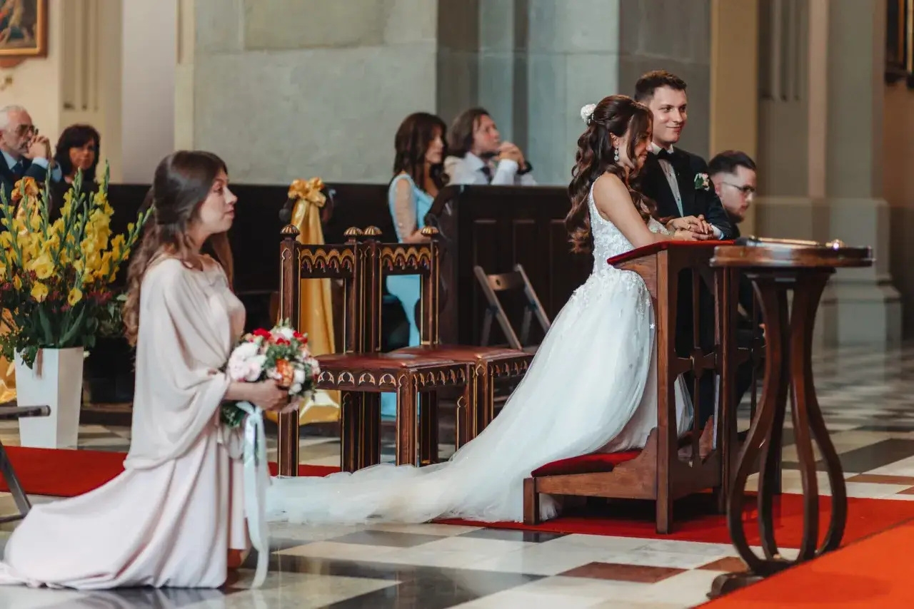 Ślub w kościele. Panna młoda w białej sukni klęczy przy ołtarzu. Obok niej świadek. Klęczy też świadkowa z bukietem kwiatów. W tle goście. Obowiązki świadkowej lista.