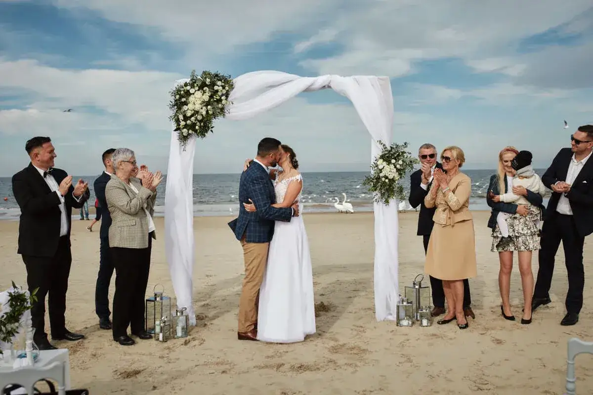 Para młoda całuje się pod baldachimem podczas ślubu na plaży w Polsce. Goście klaszczą, a na morzu widać łabędzie.