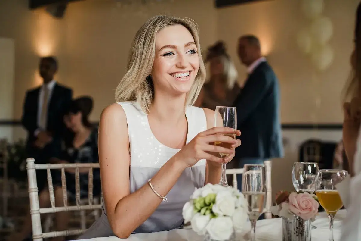 Uśmiechnięta blondynka z kieliszkiem szampana, szuka partnera na wesele. W tle goście weselni.