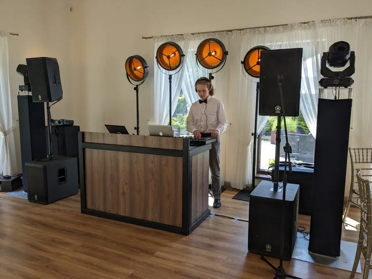 Młody DJ w muszce przygotowuje muzykę na wesele, otoczony sprzętem nagłośnieniowym i oświetleniem.